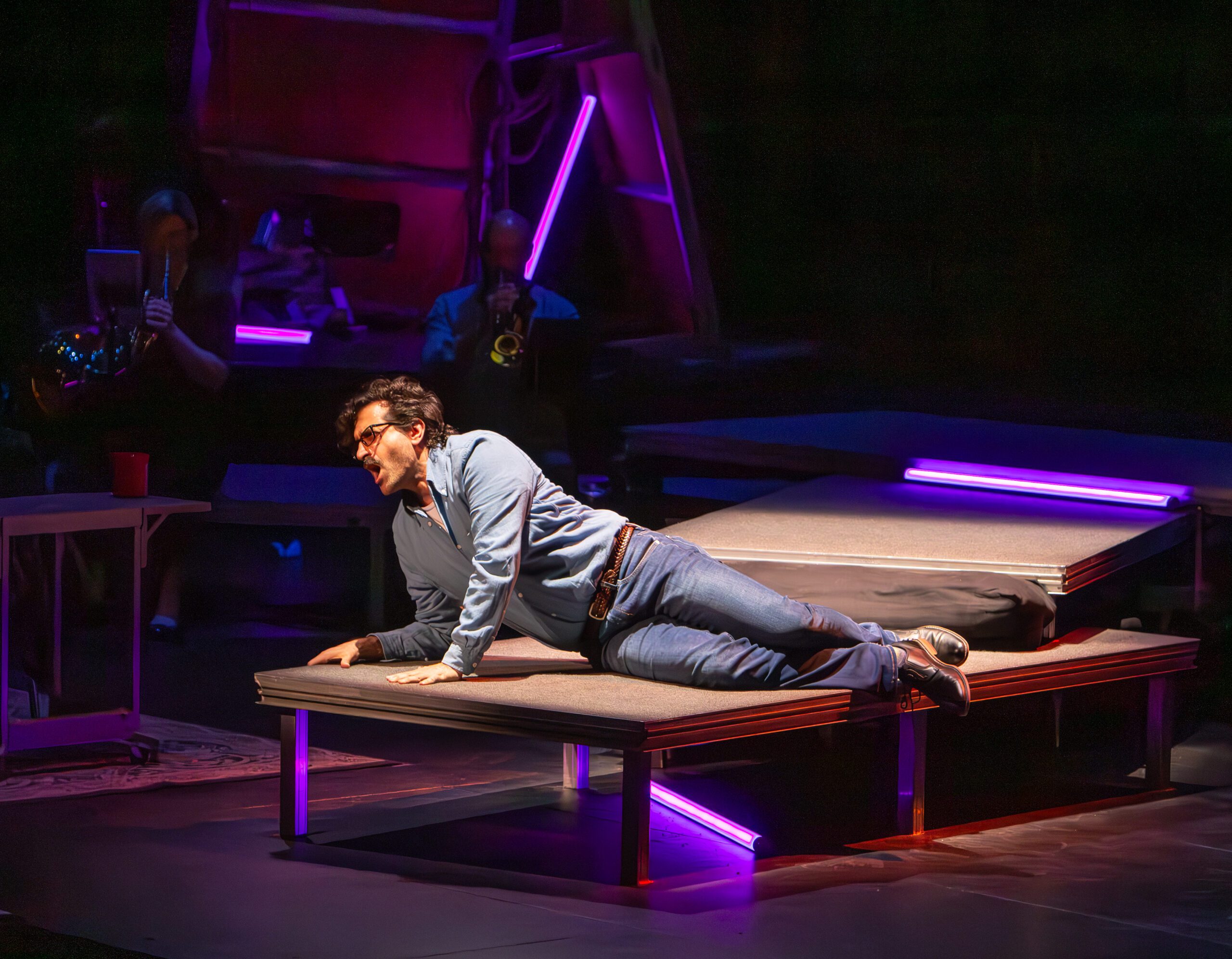 A man in glasses and casual clothes lies on a raised platform, singing passionately. Musicians play instruments in the dimly lit background with purple neon lights accenting the stage.