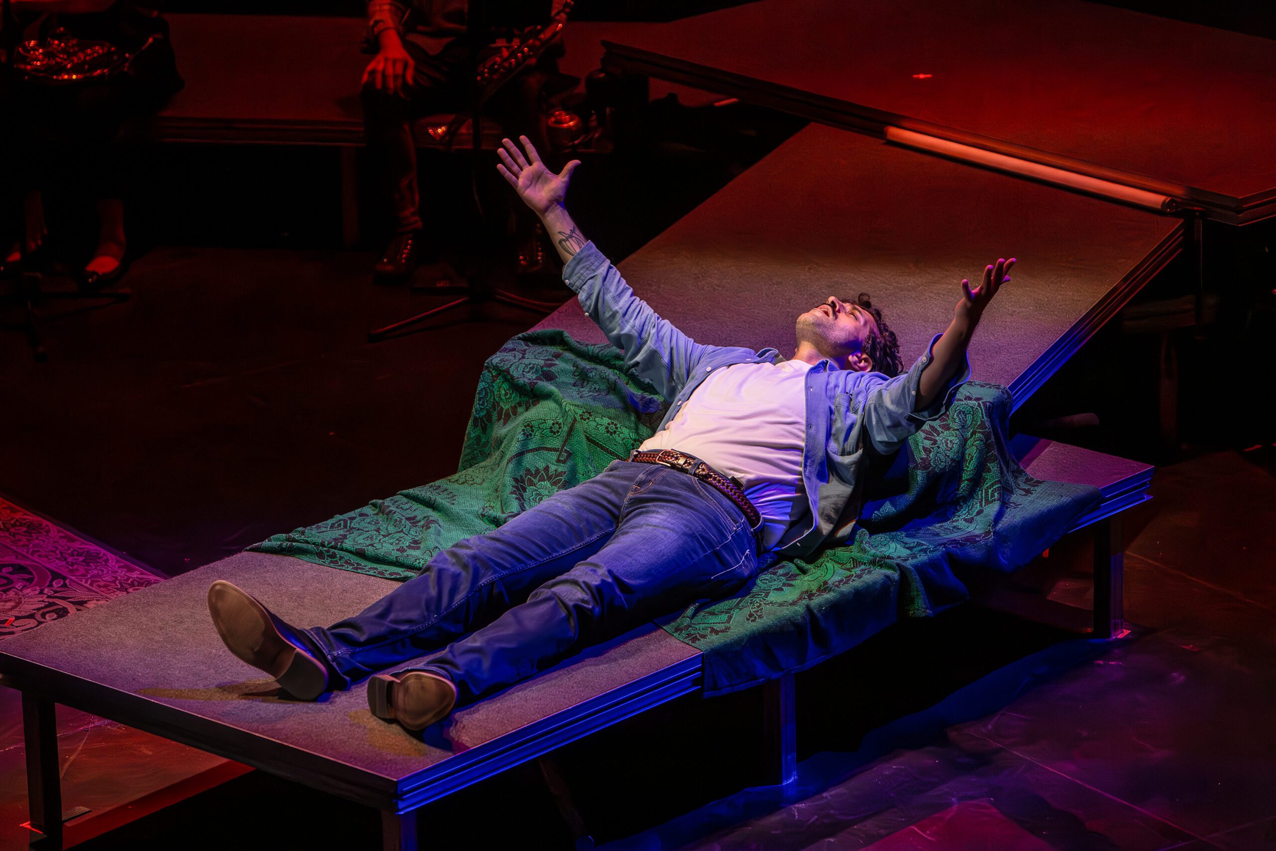 A person in jeans, a white shirt, and an open jacket lies on a platform with arms outstretched, looking upward under dramatic stage lighting.