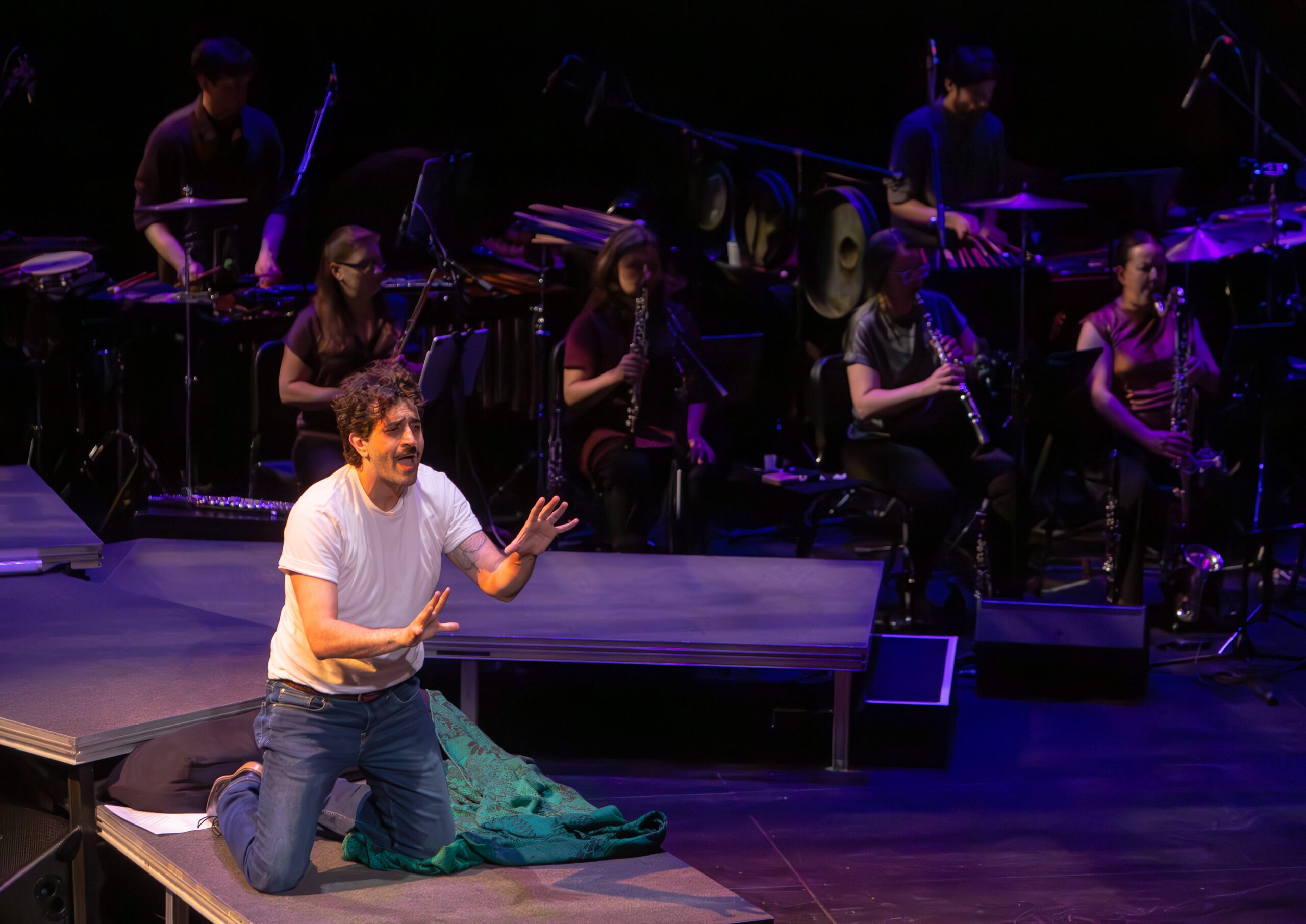 A man in a white shirt and jeans kneels on stage, gesturing emotionally. Behind him, musicians play various instruments under stage lights, creating a dramatic performance scene.
