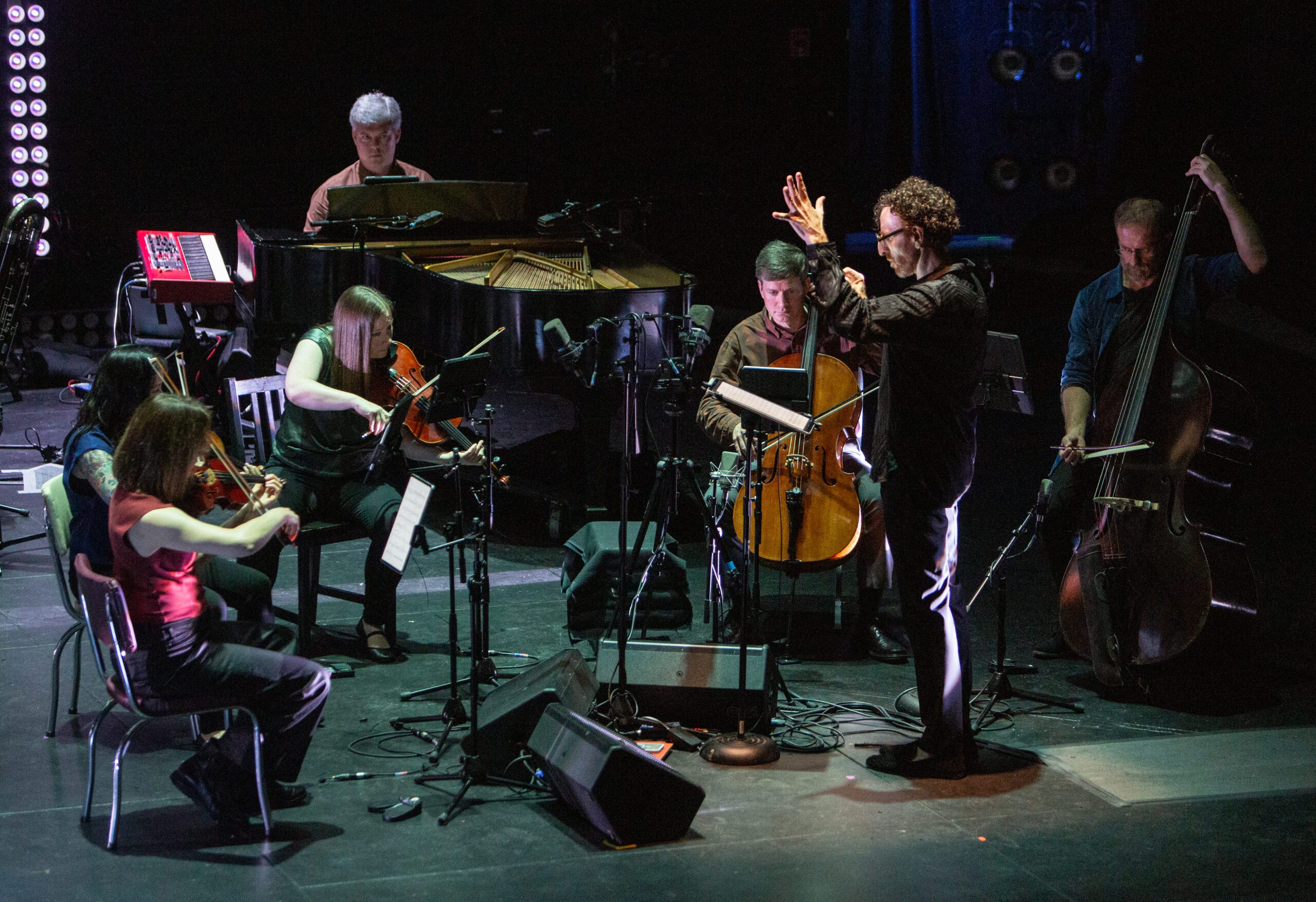 A conductor leads a small ensemble with strings and a pianist on a dimly lit stage. Musicians are seated with music stands, and various instruments, microphones, and equipment surround them.