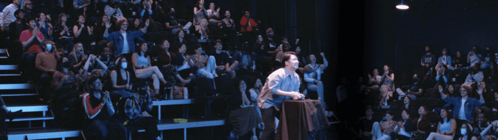 A person stands at a podium on stage facing a large, diverse audience seated in bleachers, many of whom are clapping or watching attentively in a dark theater.