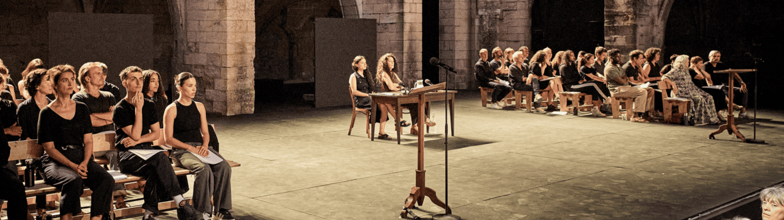 A group of people dressed in black sit on wooden chairs in rows on a stage, some applauding, with two people seated at desks facing the audience under stone archways.