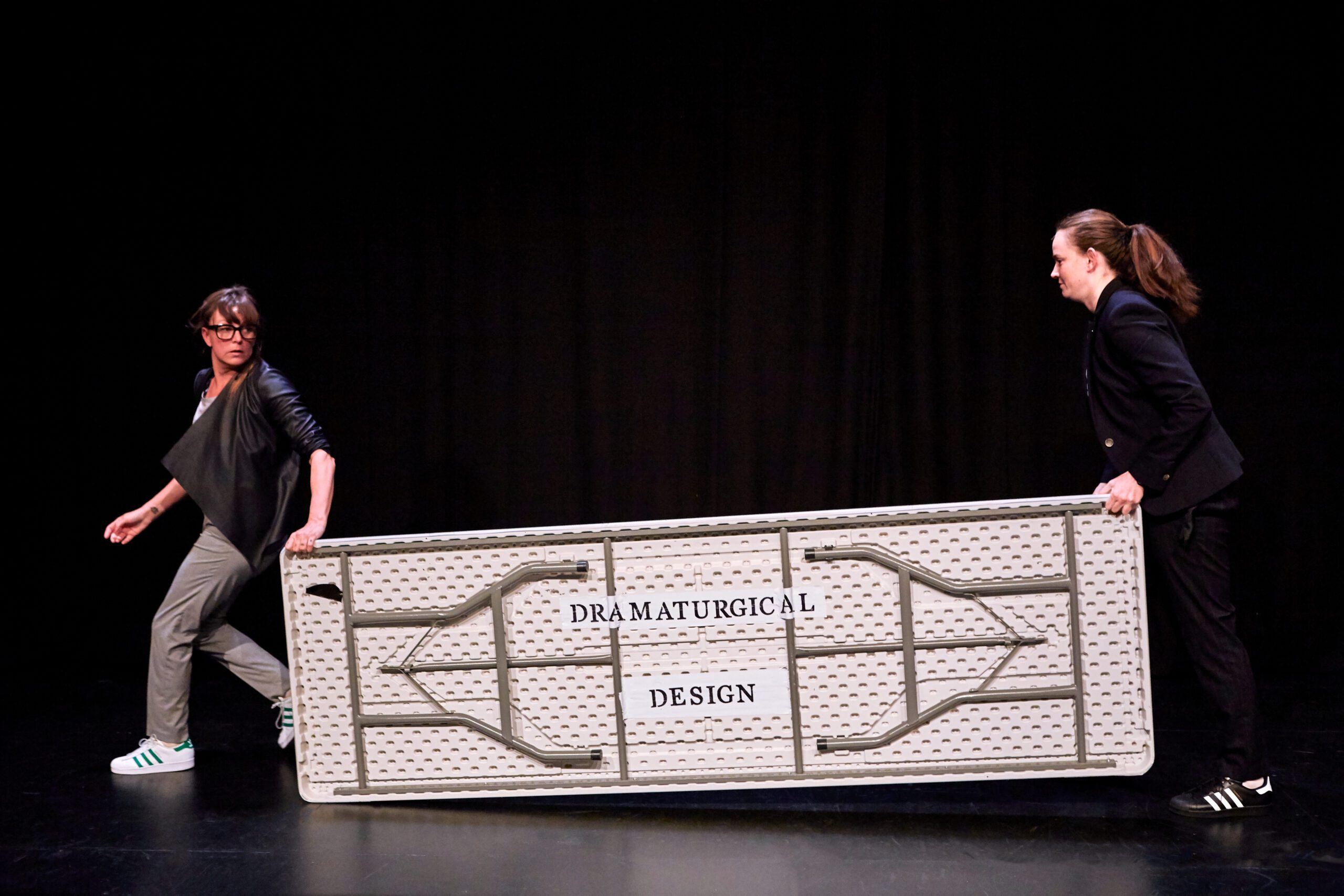 Two people in dark clothing lift and carry a large, gray folding table labeled “DRAMATURGICAL DESIGN” on a black stage.