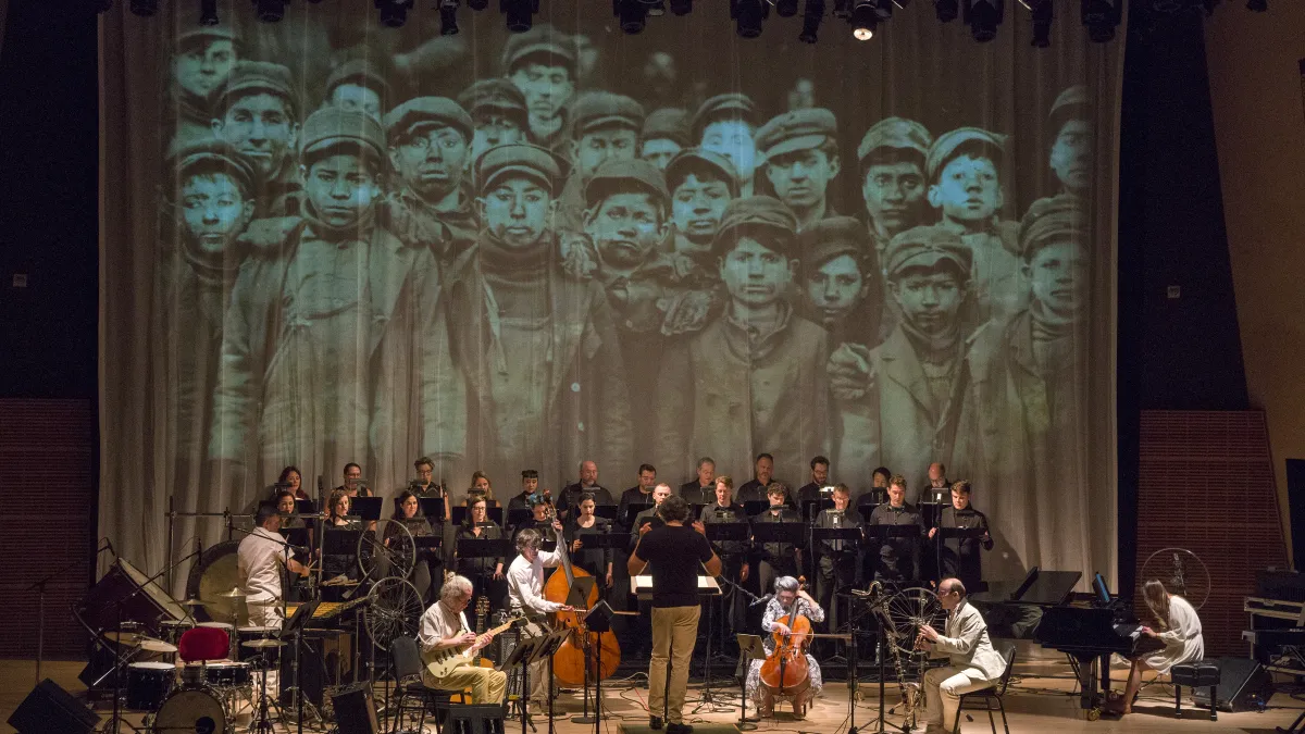 A choir and musicians perform on stage, with a large historic black-and-white photo of children in hats and coats projected behind them.