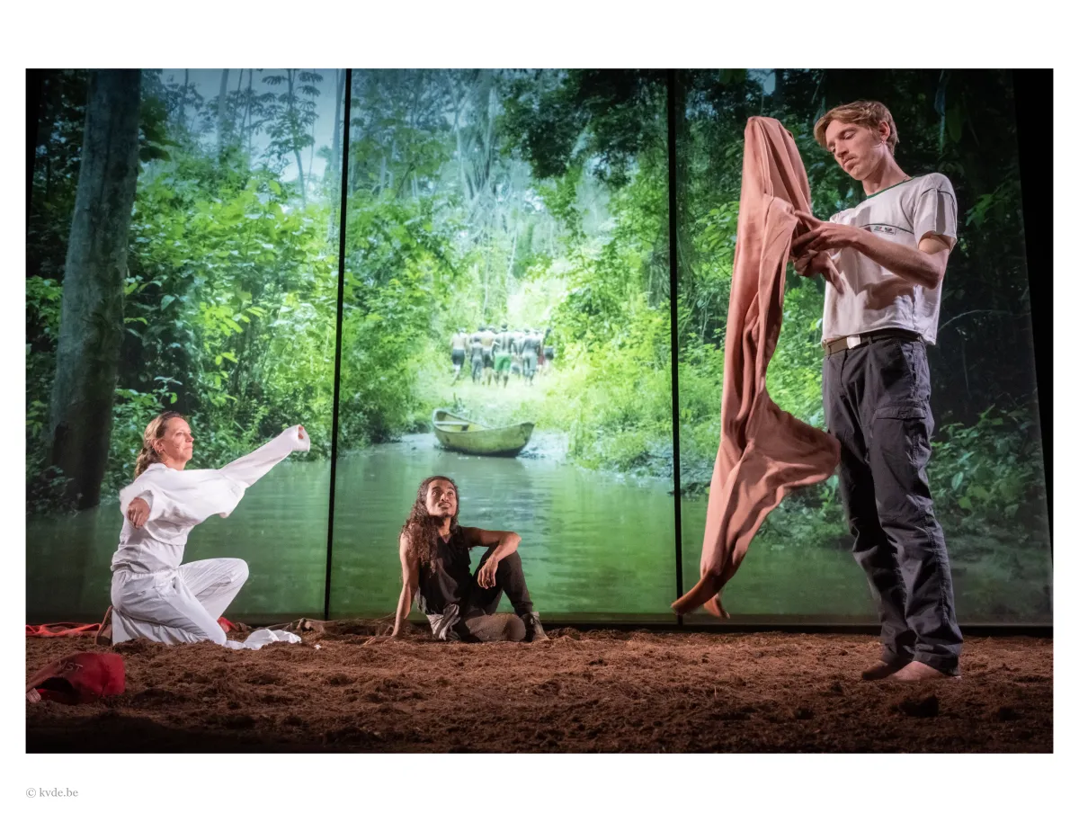 Three people perform on a dirt-covered stage with a forest and river backdrop. One kneels, holding white cloth; another sits on the ground; the third stands, unfolding brown fabric. A boat is visible in the forest scene behind them.
