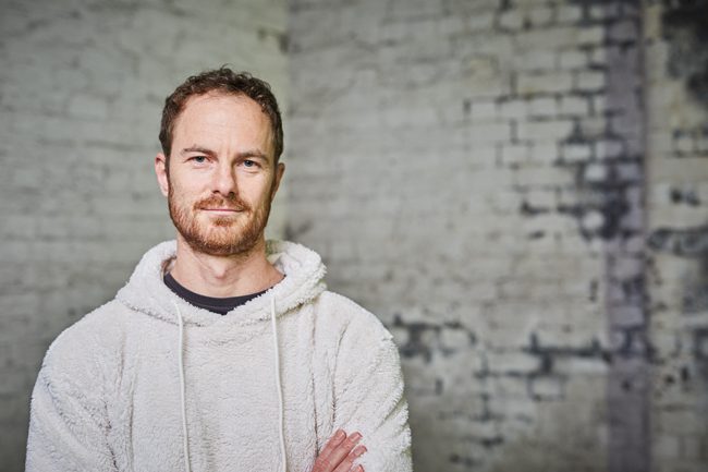 A man with short curly hair and a beard, wearing a light-colored fleece hoodie, stands with arms crossed in front of a white brick wall.