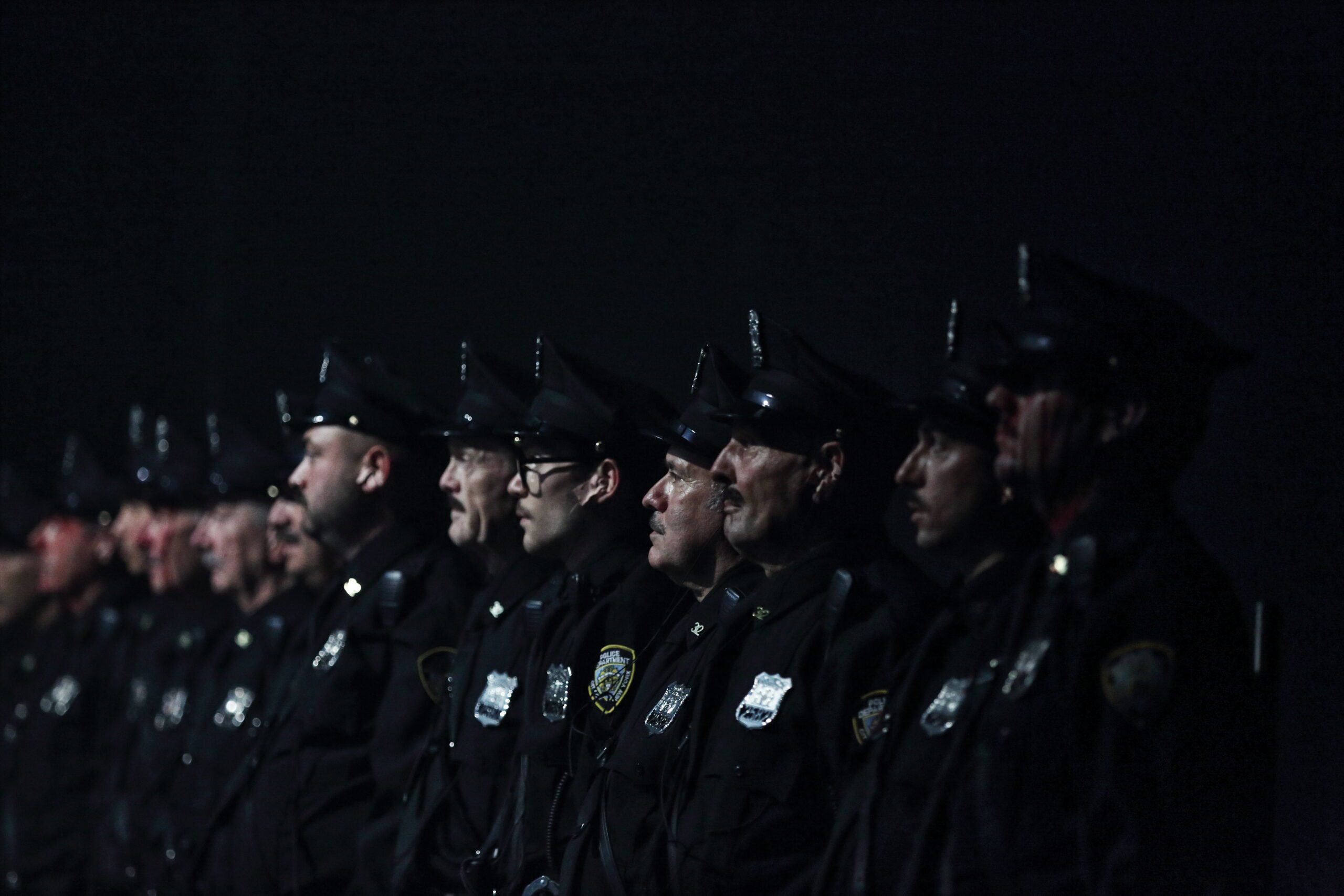 A row of police officers in uniform stand at attention in a dimly lit setting, facing forward.