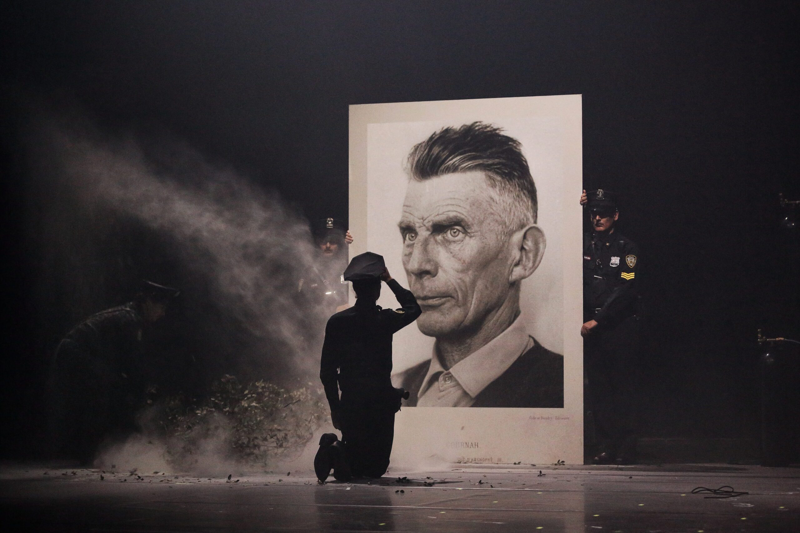 A person kneels and salutes in front of a large portrait of a man, while two uniformed officers stand beside the portrait on a dimly lit, smoky stage.