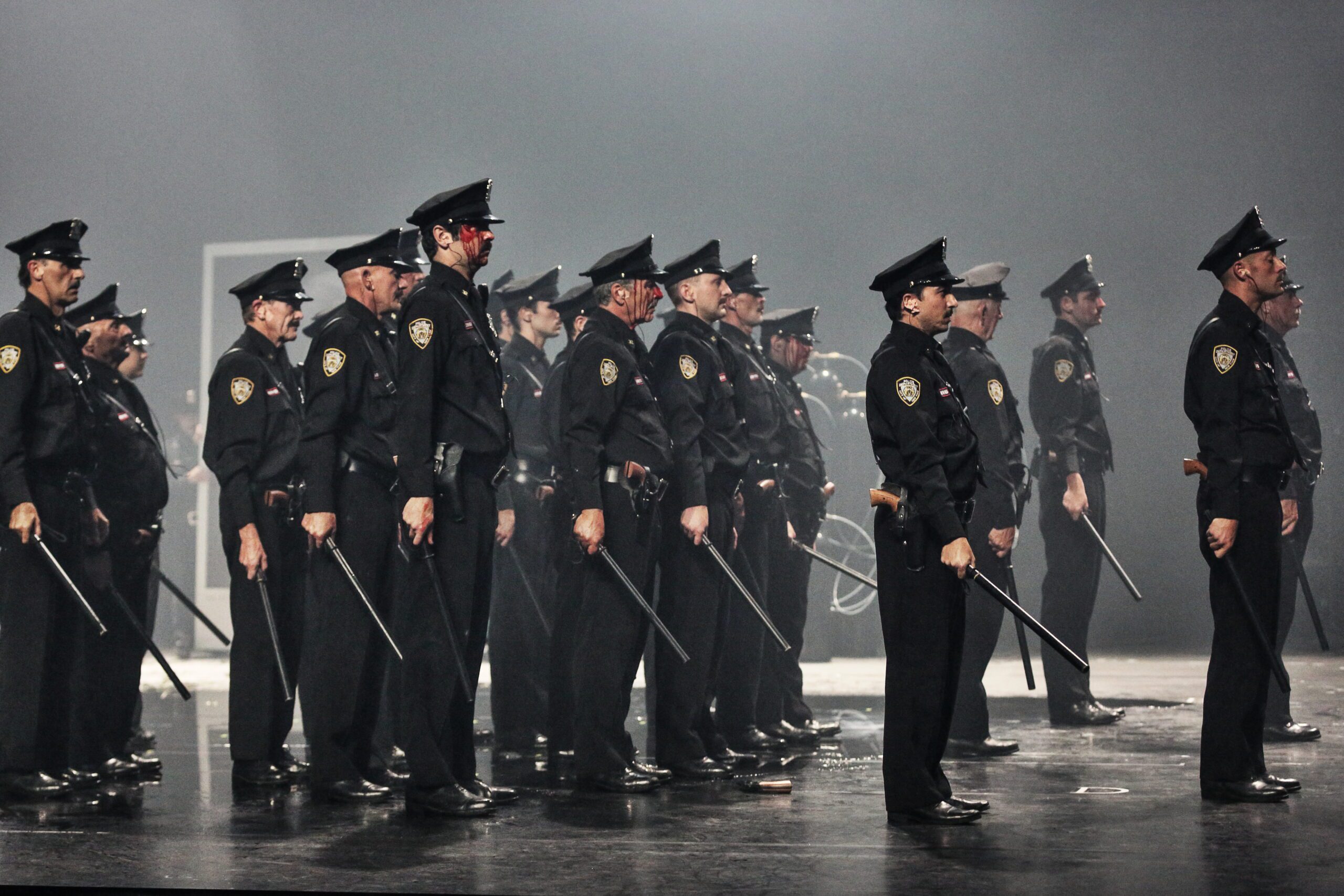 A group of police officers in uniform stand in formation on a stage with batons at their sides, facing forward under dramatic lighting.