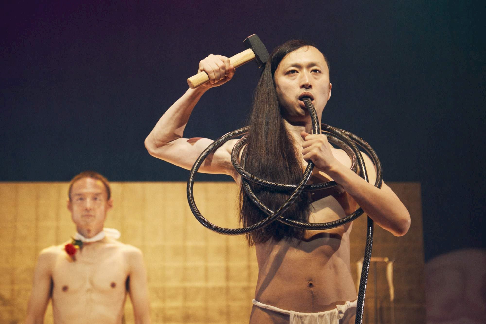 Two people stand on stage with minimal clothing; the person front holds a hammer and has a vacuum hose wrapped around their body, speaking into a microphone, while another stands in the background with a flower at their neck.