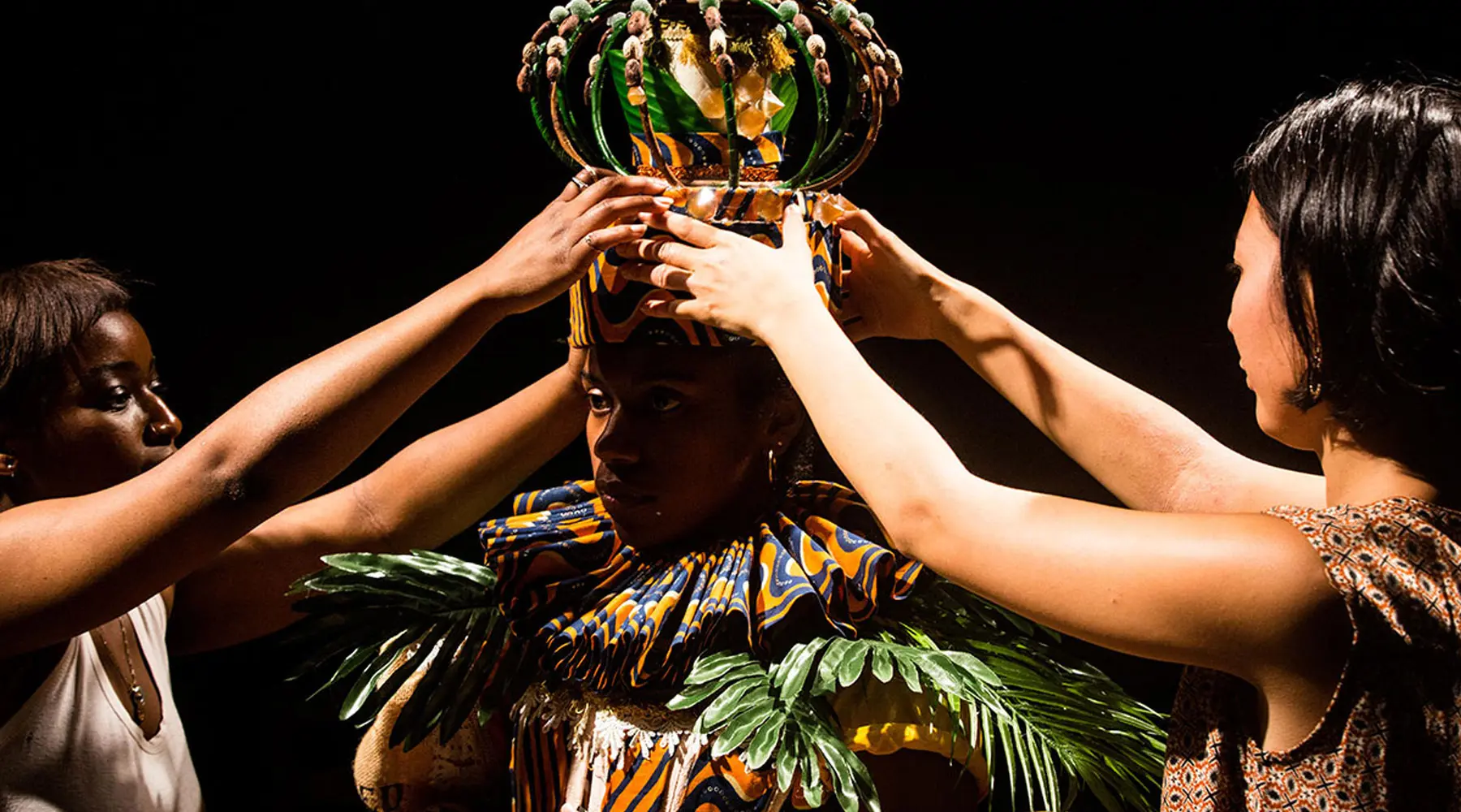 Three people place an elaborate, colorful crown on a person dressed in vibrant, patterned clothing with green leaf-like details, against a dark background.