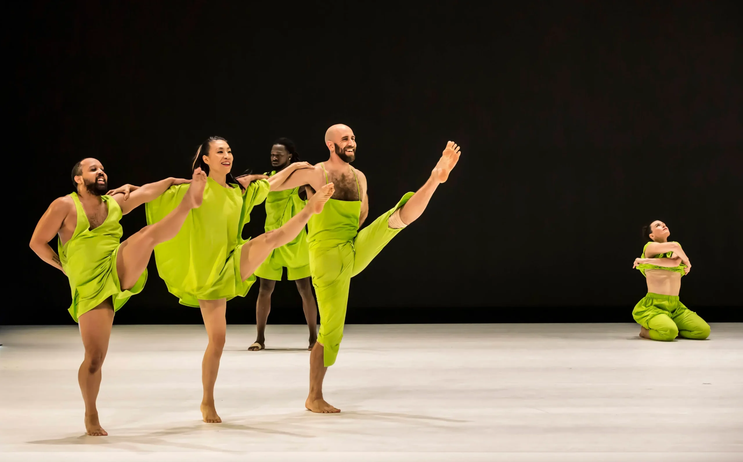 Five dancers in bright green costumes perform on stage; three extend their legs high, while one hugs their knees and another stands in the background. The stage background is dark, highlighting the dancers’ movements.