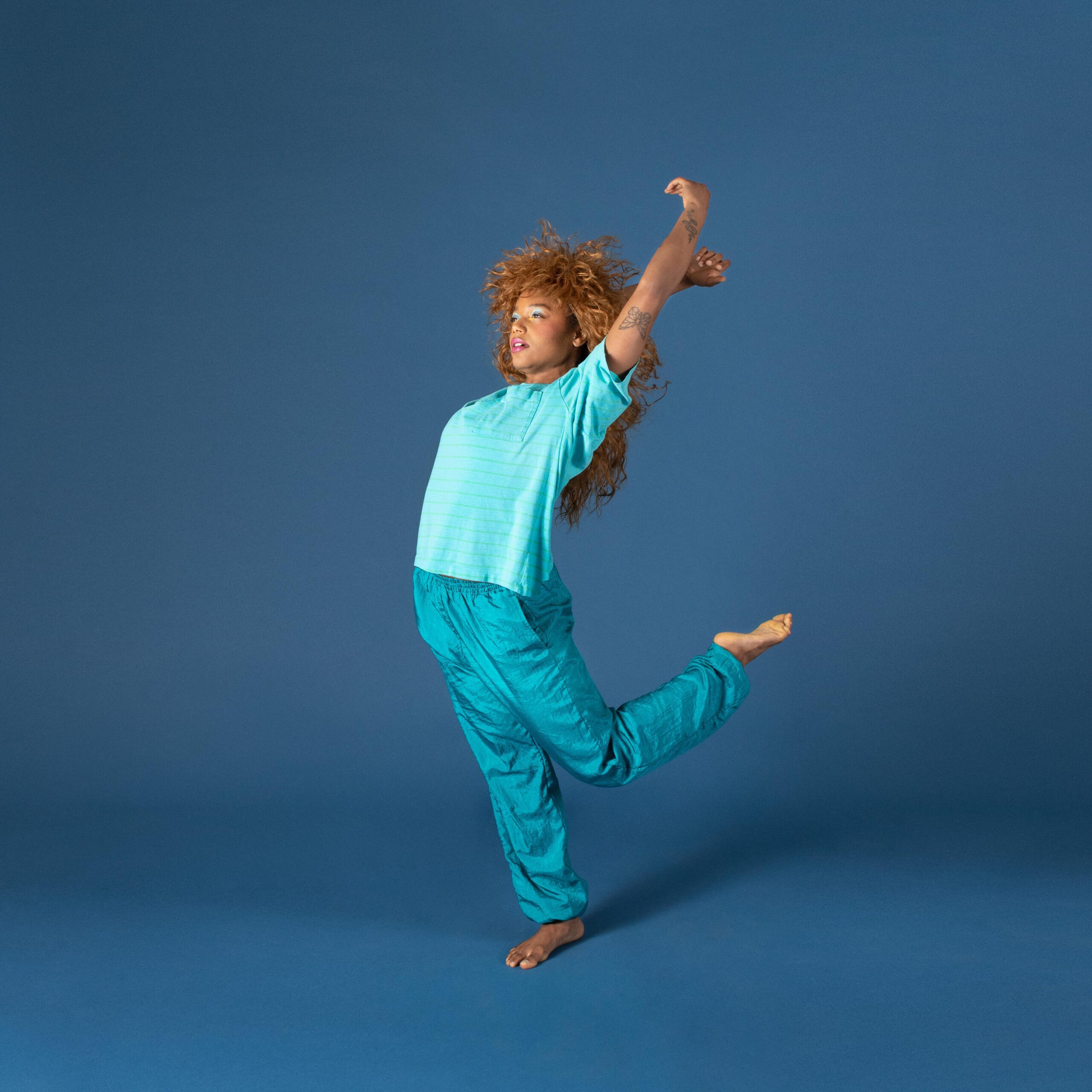 A person with long curly hair, wearing a turquoise shirt and pants, poses mid-leap against a solid blue background with one leg bent and arms raised above their head.