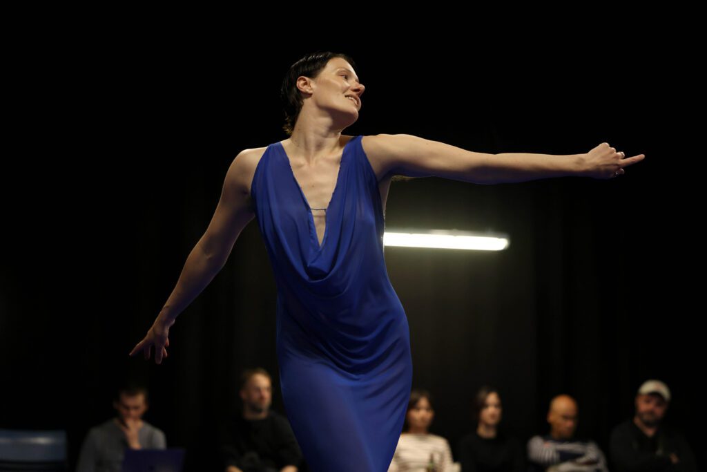 A dancer in a blue dress performs gracefully on stage, extending one arm outward. Several people are seated in the background, watching attentively.