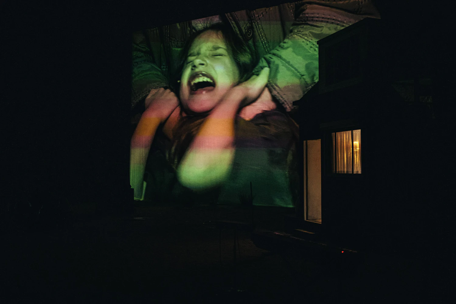 A large, distressed image of a screaming child is projected onto a dark wall at night; a small lit window appears on the right side of the scene.