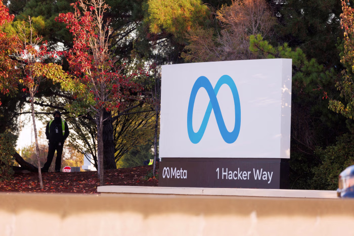 A sign with the Meta logo and text “Meta 1 Hacker Way” stands outdoors near trees with autumn leaves. A person is visible in the background under the trees.