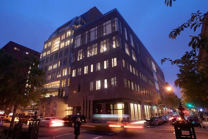 A modern building with illuminated windows at dusk, located on a busy city street with cars and trees visible in the foreground.