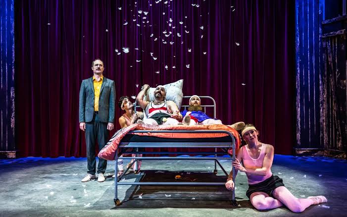 Five actors perform on stage around a hospital bed with red curtains behind them; feathers float in the air, creating a dramatic and surreal scene.