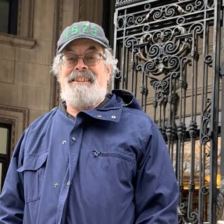 An older man with a gray beard and glasses, wearing a dark blue jacket and a cap that says “1973,” stands in front of an ornate iron gate and stone building.