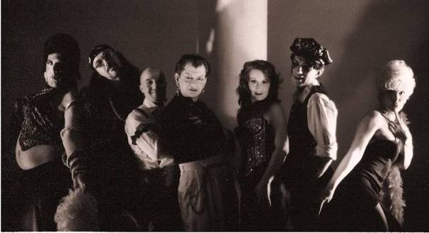 Seven people in dramatic costumes pose together in black and white, with theatrical lighting creating strong shadows and a vintage, cabaret-style atmosphere.