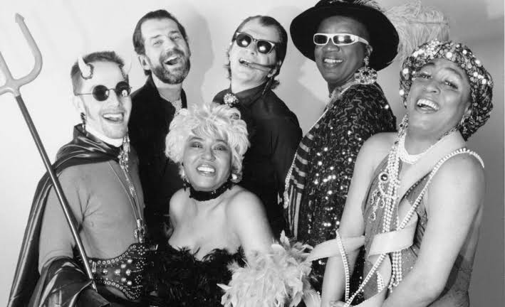 A group of seven people in costumes and accessories, smiling and laughing together. Some wear sunglasses, hats, sequins, and feather boas; one holds a trident. The photo is black and white.