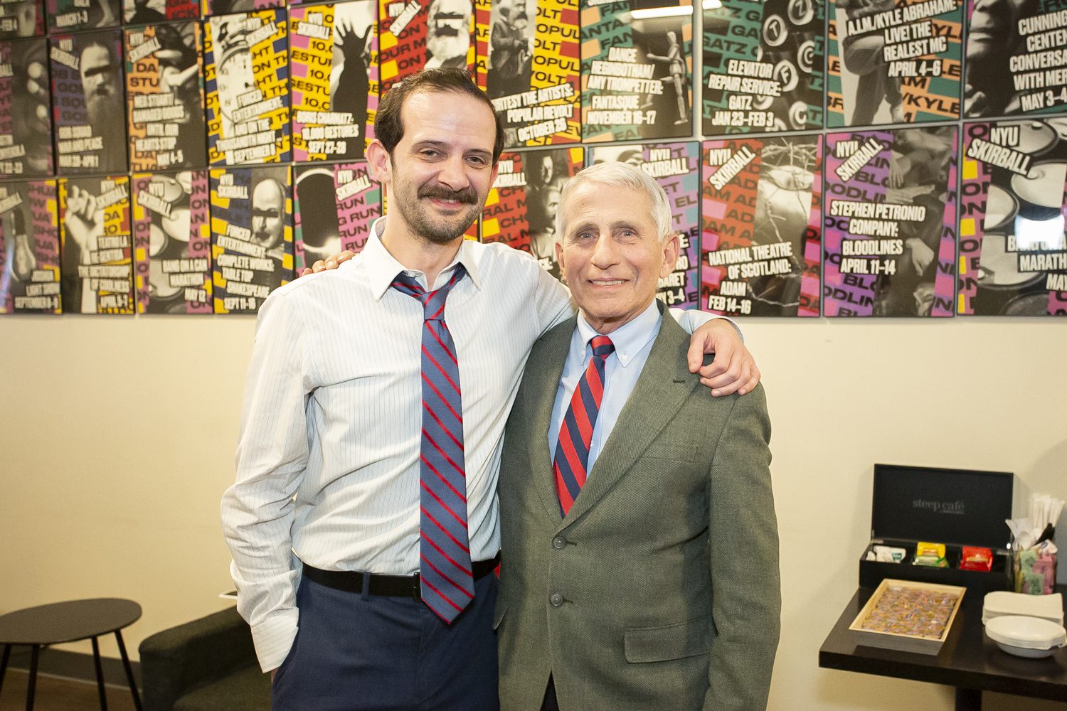 Two men in shirts and ties stand smiling with arms around each other in front of a colorful wall covered with various posters. A small table with tea, coffee, and supplies is visible to their right.