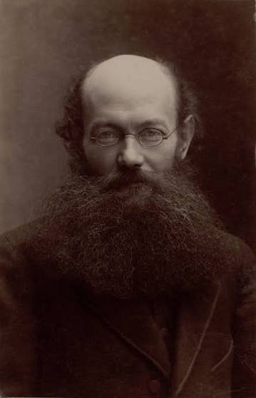 A sepia-toned portrait of a man with a bald head, round glasses, and a long, thick beard. He is wearing a dark suit and looks directly at the camera.