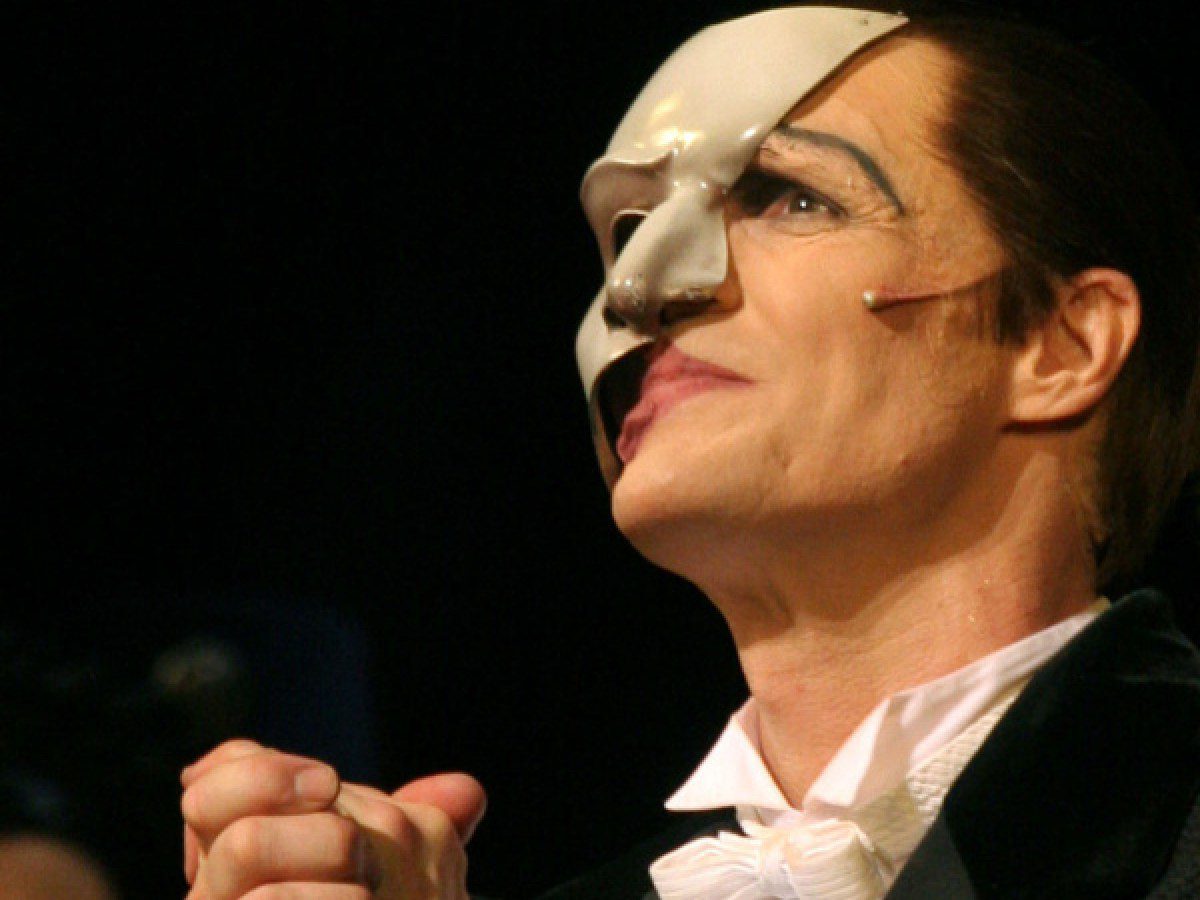 A person in formal attire wears a white half-face mask, covering the right side of their face. They have a visible microphone and hands clasped, looking upward, possibly performing on stage.