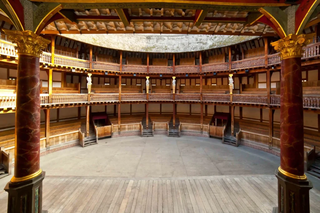 A view of an empty, circular wooden theater with three balcony levels, ornate pillars, wooden benches, and an open stage floor. Warm sunlight highlights the detailed architecture.
