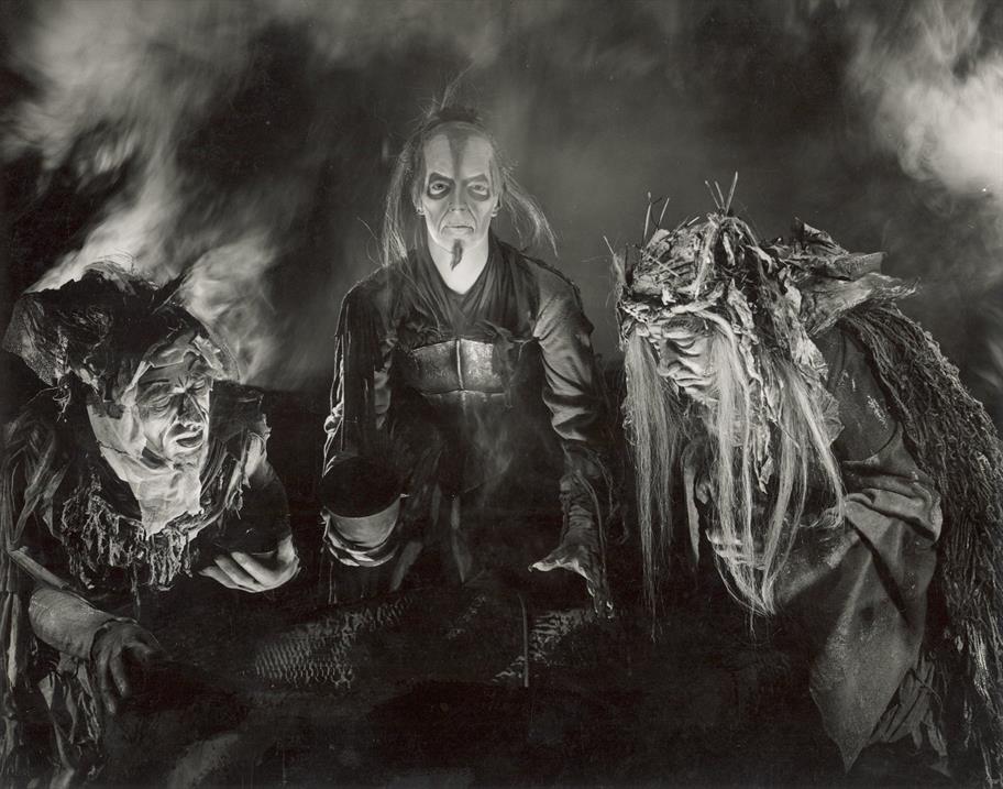 Three people in eerie costumes and makeup, resembling witches, gather around a table in a smoky, dark setting. Their expressions are intense and dramatic, and the atmosphere feels mystical and ominous.