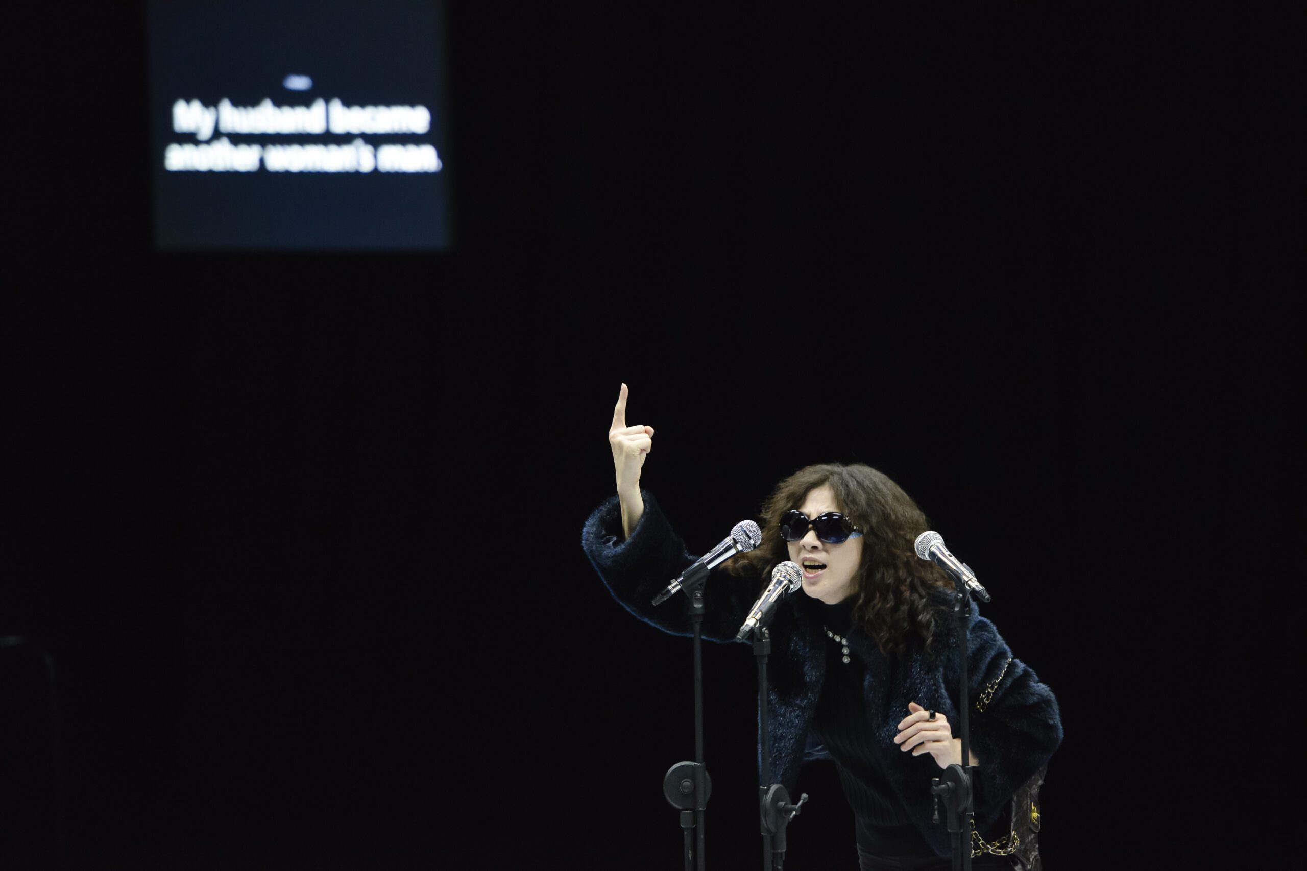 A woman in dark clothes and sunglasses speaks passionately into two microphones, raising one finger. In the background, a screen displays the text: “My husband became another woman’s man.”.