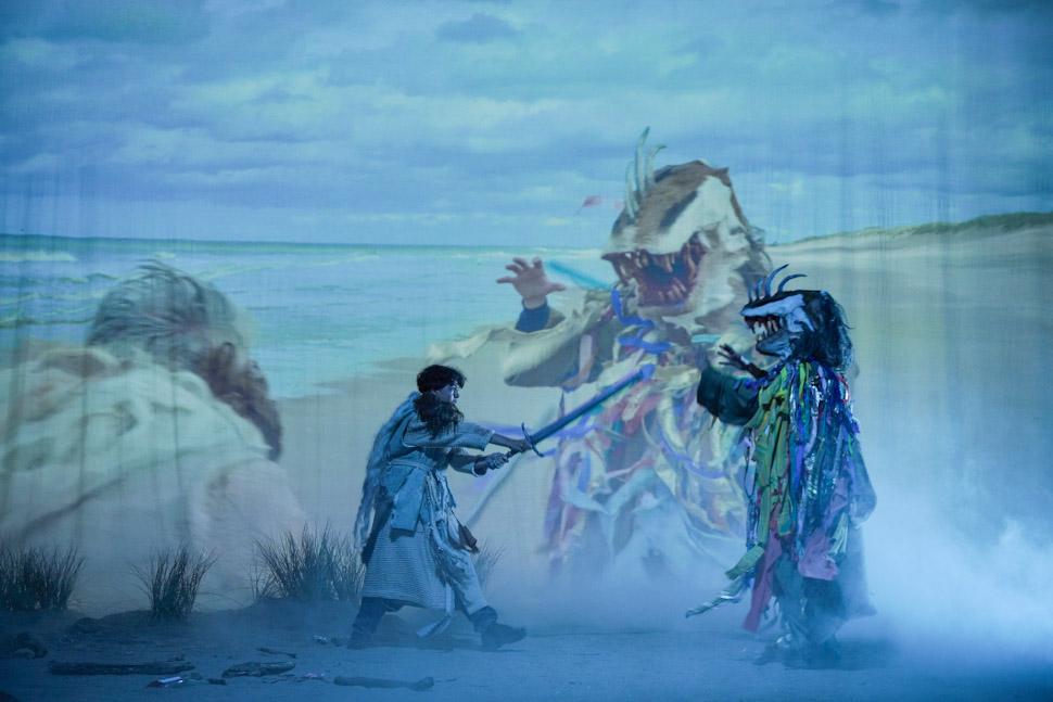 Two people in colorful, elaborate costumes face off with swords on a misty stage, with a beach scene projected behind them showing giant monster figures. Dune grass lines the front of the stage.