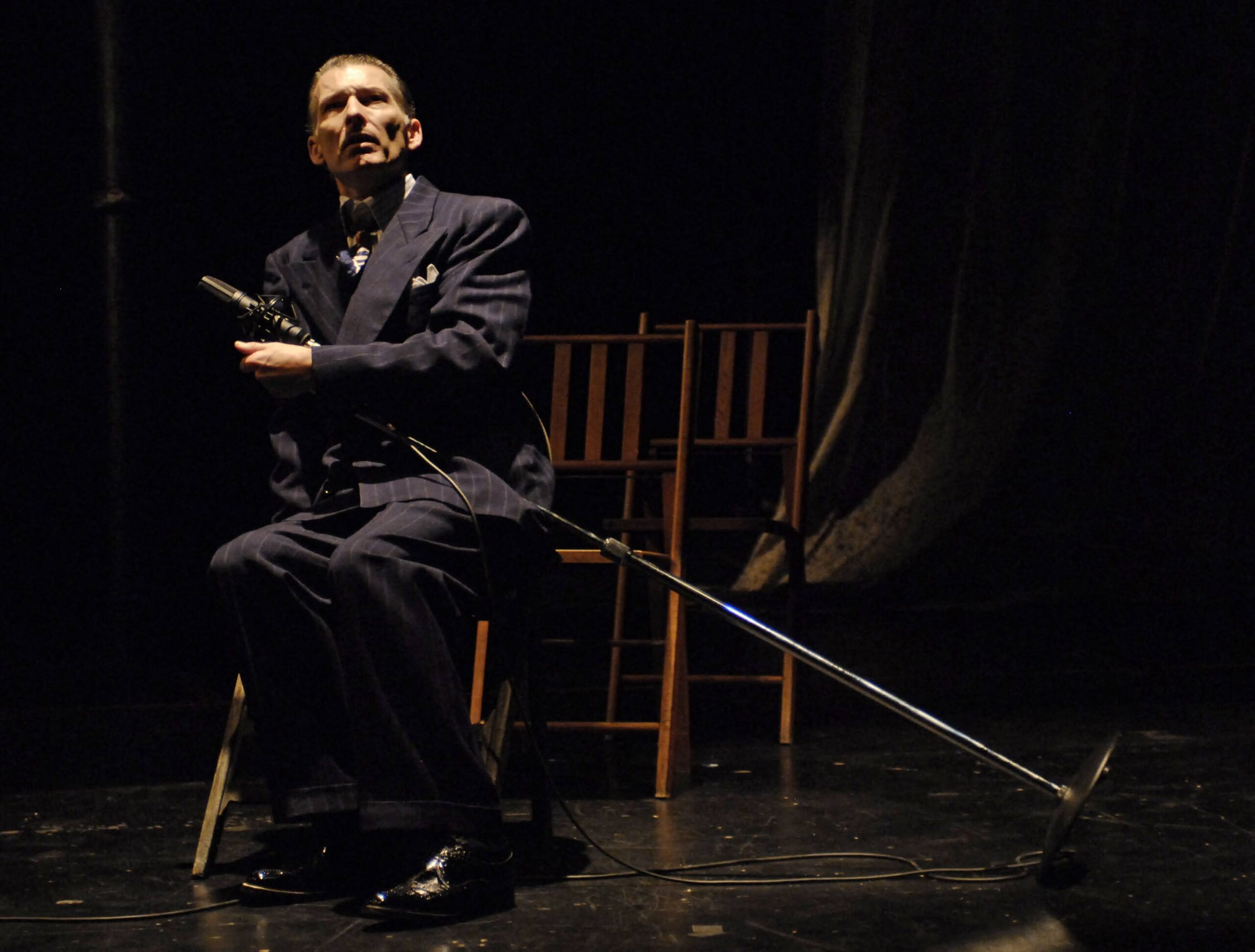 An older man in a dark suit sits on a wooden chair onstage, holding a microphone and stand. The background is dimly lit with empty chairs and draped fabric.