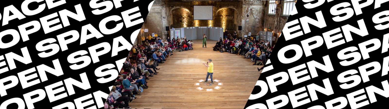 A person stands speaking in the center of a large wooden hall, surrounded by seated people in a circle. The words “OPEN SPACE” repeat in bold, white text on a black background along the sides.