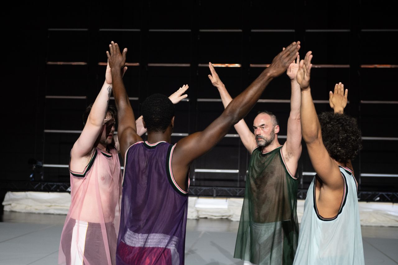 Four people in sleeveless mesh shirts stand in a small circle, raising their arms and touching hands above their heads, as if in a group cheer or celebration. The background is dark and minimalist.