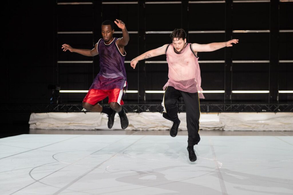 Two dancers in loose tops and athletic pants leap mid-air on a lit stage with a white floor and dark background, their shadows visible beneath them.