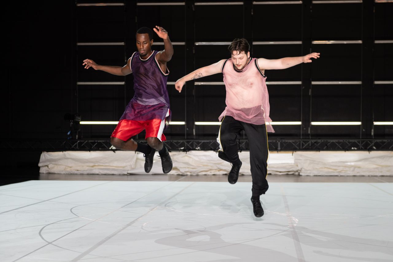 Two dancers in loose tops and athletic pants leap mid-air on a lit stage with a white floor and dark background, their shadows visible beneath them.