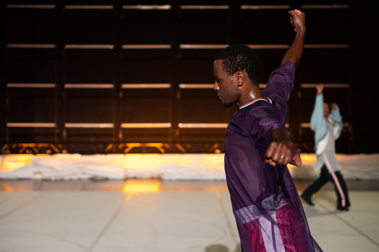 A person in a purple shirt dances with one arm raised, while another dancer in the background also lifts an arm. The setting is dimly lit, with a dark backdrop and warm lights on the floor.