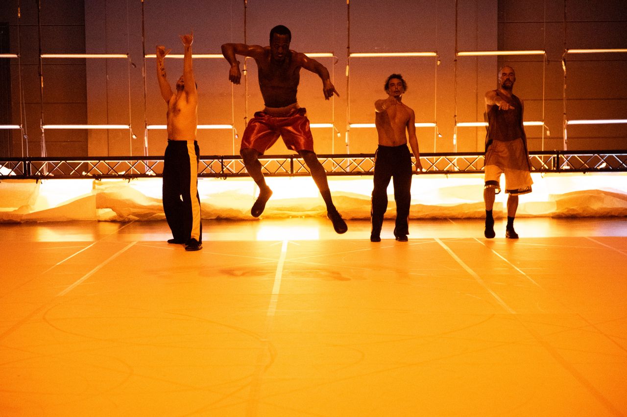 Four men perform on a brightly lit stage; the second from the left is mid-jump, while the others stand or raise arms. Warm lights and geometric wall patterns create a dramatic backdrop.