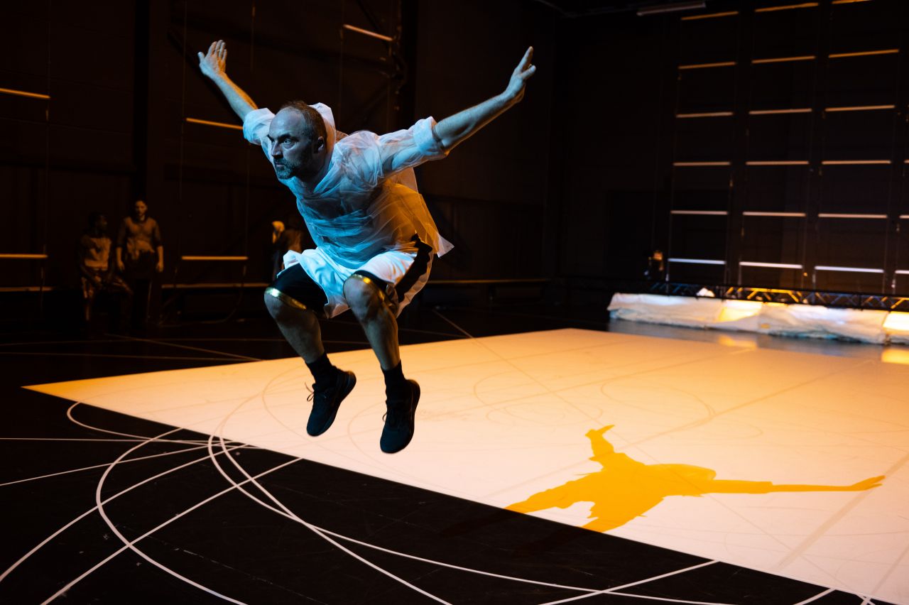 A person in a white shirt and shorts leaps mid-air on a stage, arms outstretched, with their shadow cast on a lit white floor marked with circular lines. The background is dark with faint figures and geometric patterns.