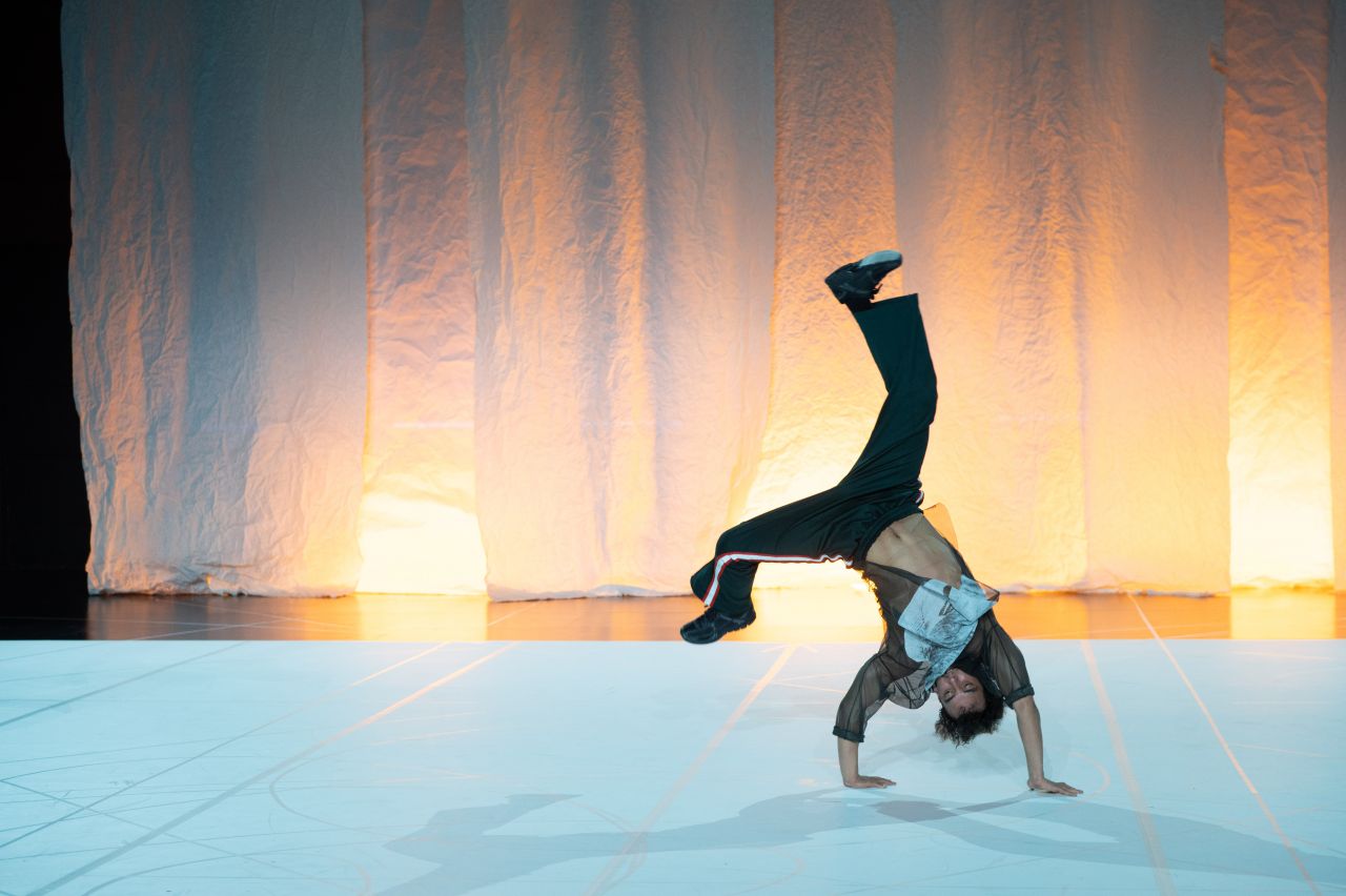 A dancer performs a one-handed handstand onstage, with legs extended and a sheer backdrop softly lit in orange tones behind them.