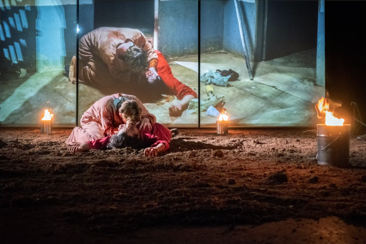 Two people in dramatic pose on a dirt-covered stage with lit torches and a metal bucket, while a large video projection of the same scene appears behind them.