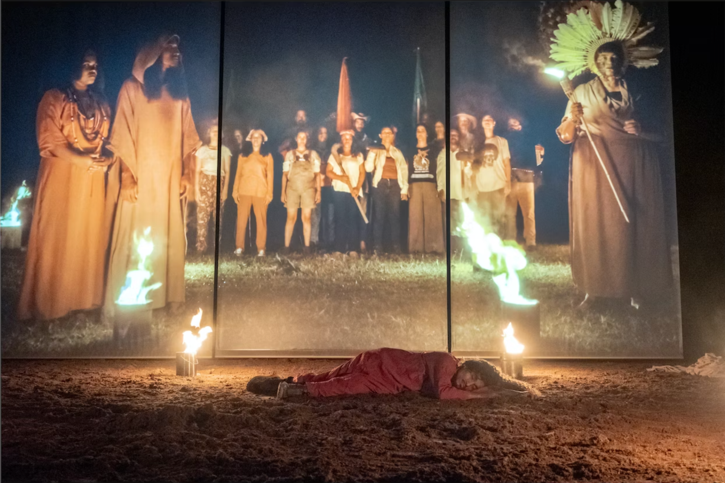 A person in red lies on sandy ground in front of a screen showing a group of people with flags and torches. Small fires burn near the person, creating a dramatic nighttime scene.