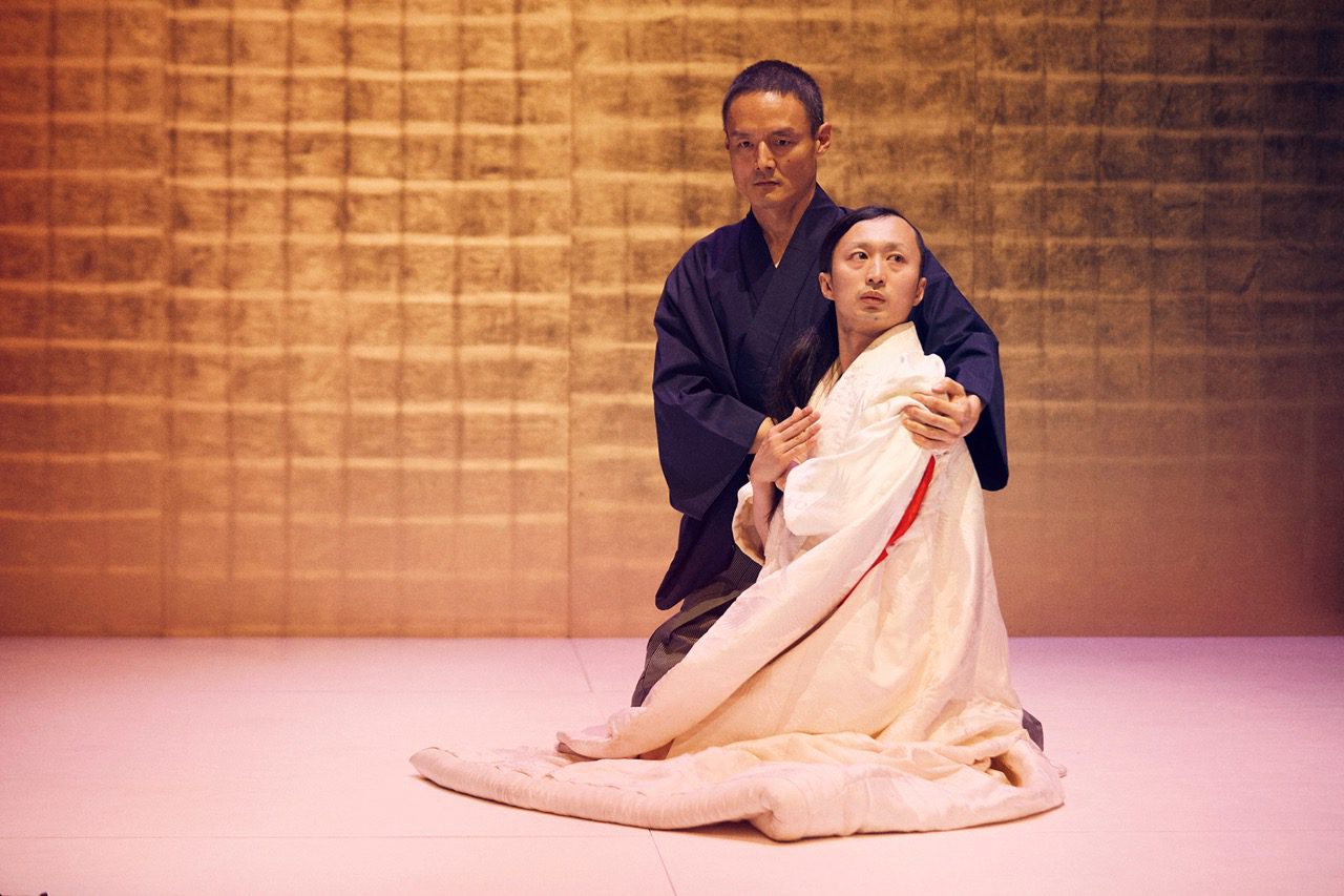 Two people in traditional Japanese attire sit on the floor against a textured golden wall; one, in dark clothing, embraces the other, who is wrapped in a white robe, both with serious expressions.