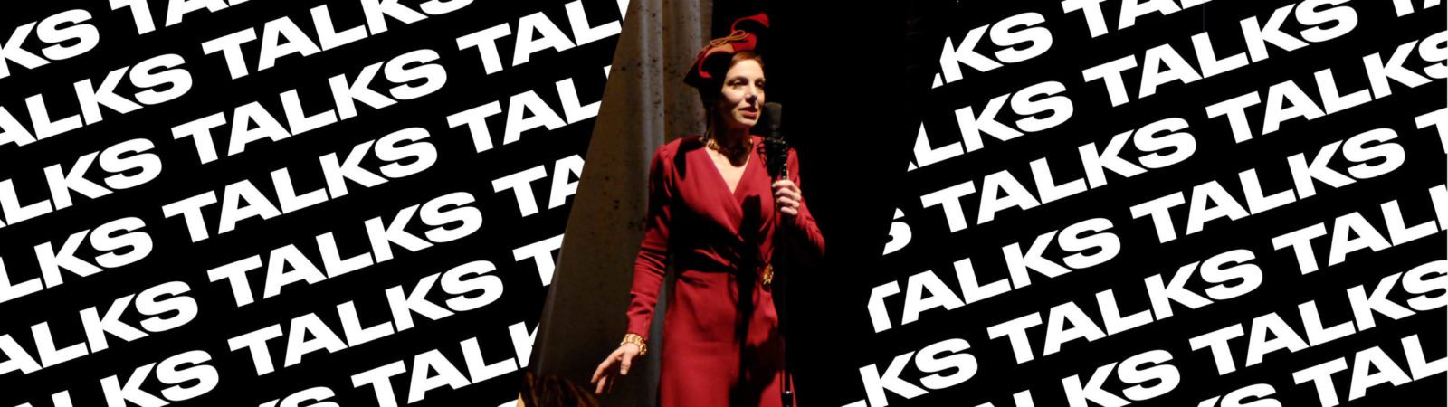 A person in a red outfit and hat speaks into a microphone onstage. The background repeats the word “TALKS” in bold, white letters on black.