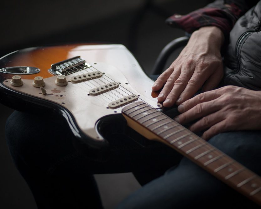 A person rests their hands on an electric guitar lying across their lap. The guitar is brown and white, and the person is wearing dark clothing.