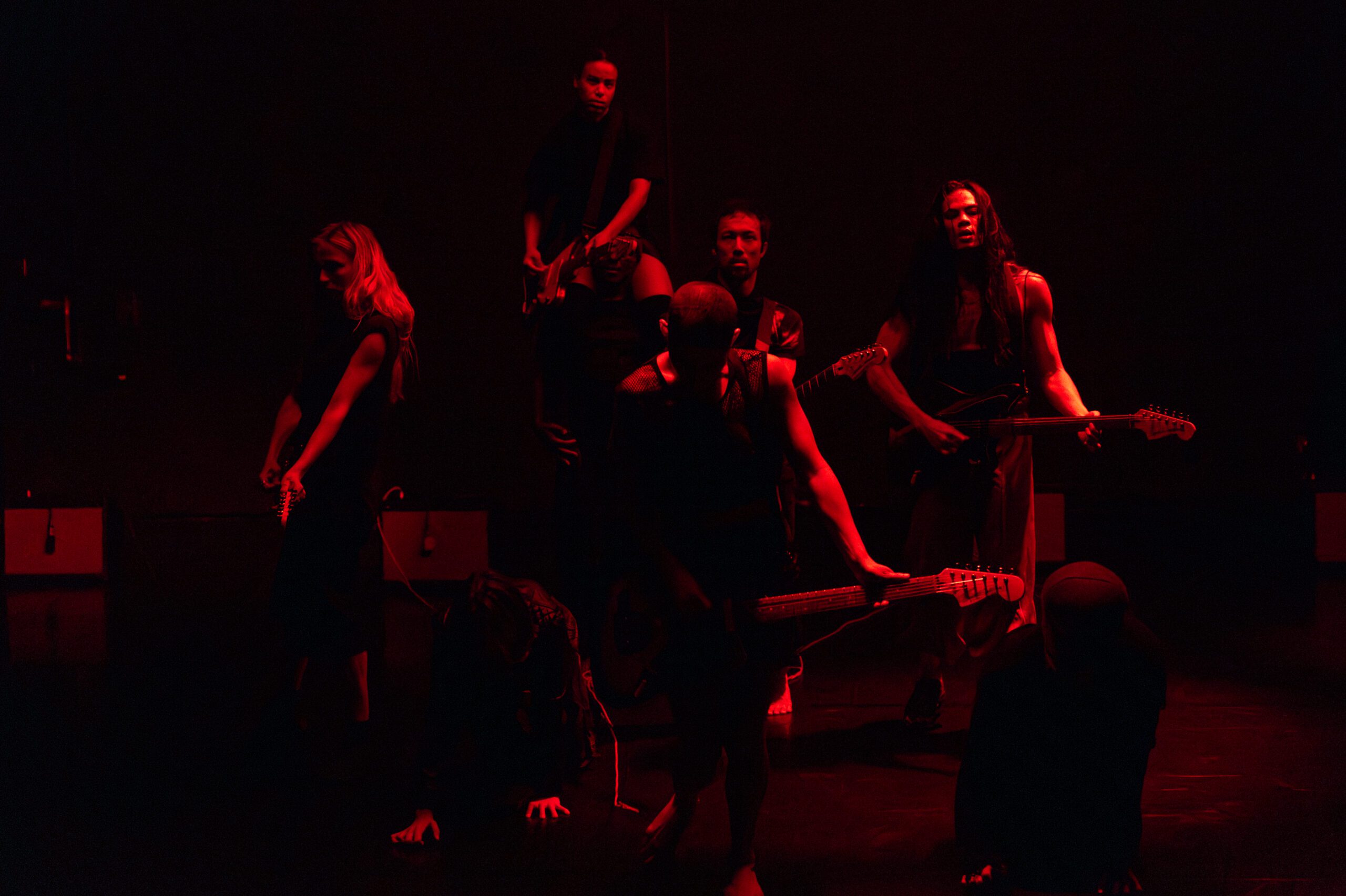 Six people holding guitars are dramatically lit in red on a dark stage, with some standing and others crouched or kneeling, creating an intense, moody atmosphere.