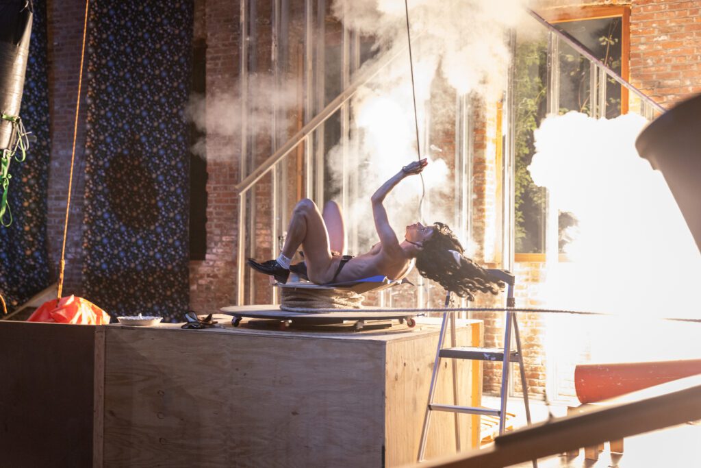 A person in a costume poses dramatically on a platform with smoke and bright light in the background, surrounded by stage props and a wooden set.