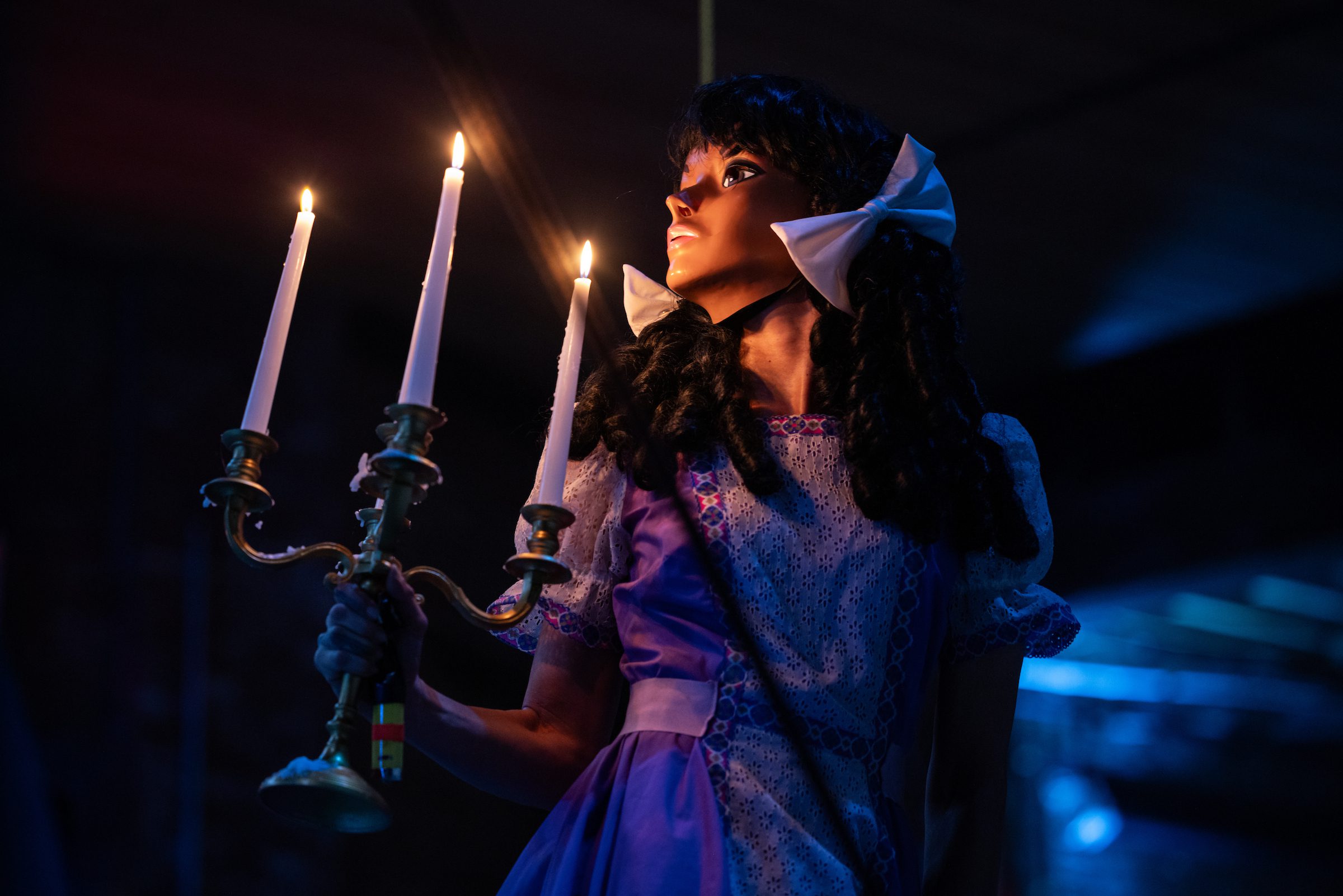 A person in a purple dress and large white bow holds a lit candelabra in a dark, moody setting, looking upward toward the candlelight.