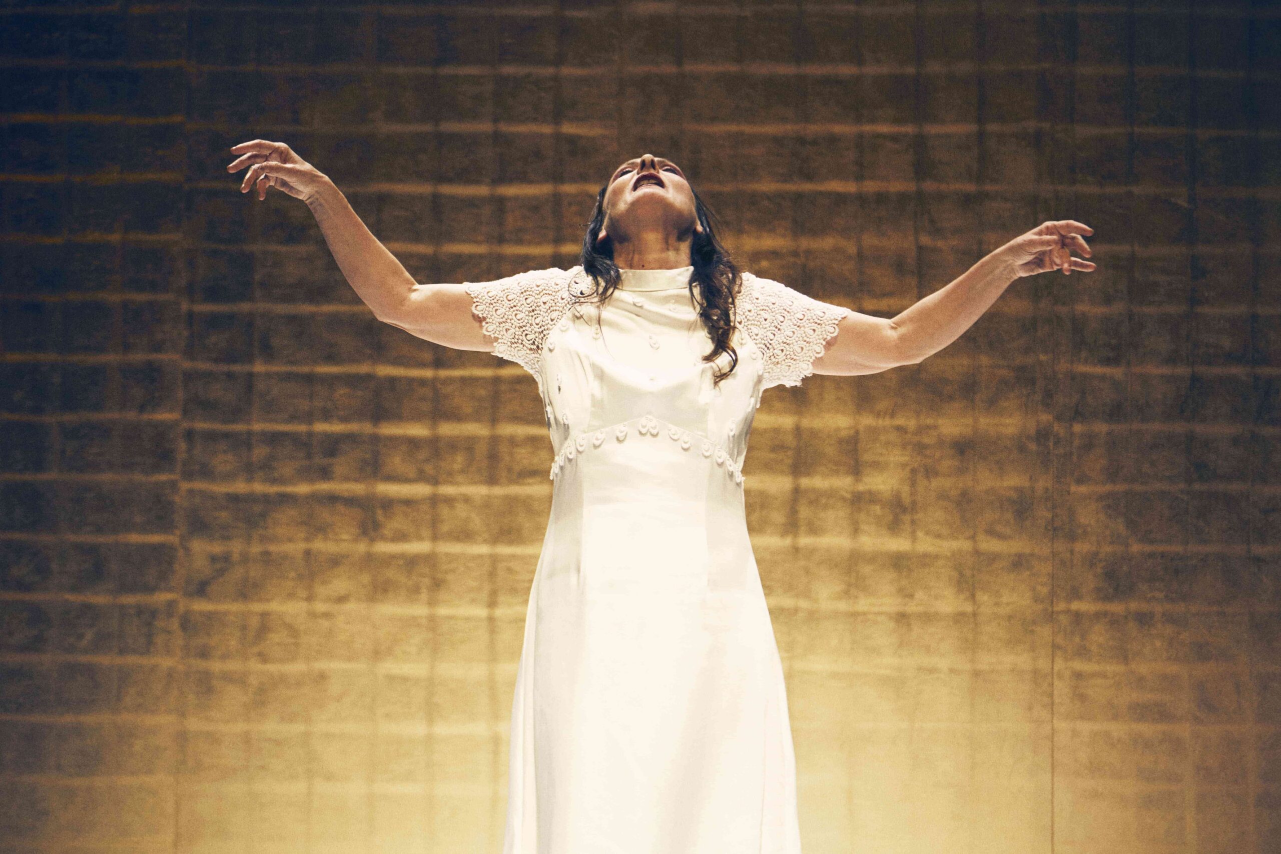 A person in a white dress stands with arms raised and head tilted back, illuminated by warm light against a textured gold background.